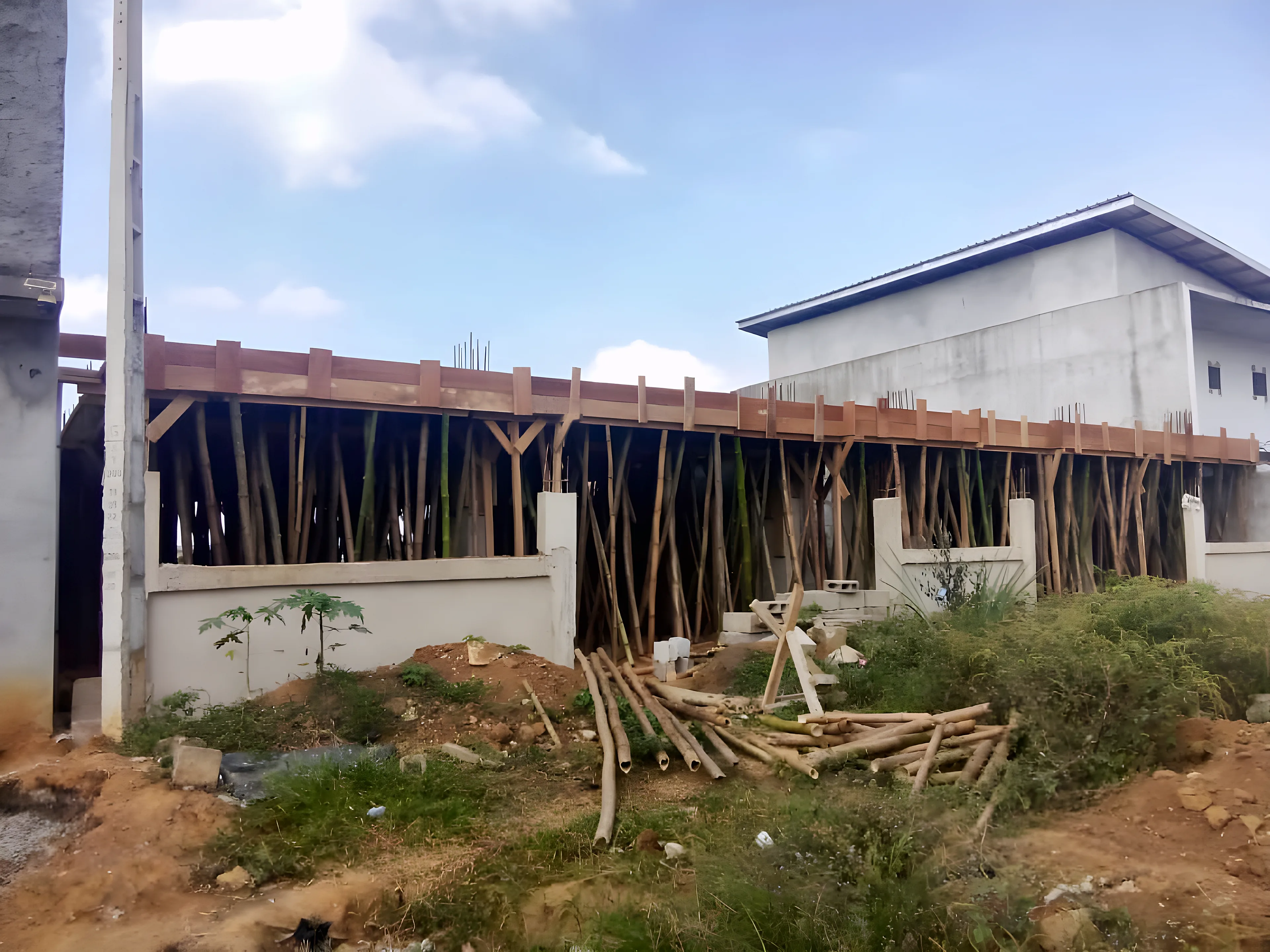Photo réelle du chantier du Bâtiment A, vue de trois quarts : coffrage bois de la dalle du 1er étage sur étais, murets du rez-de-chaussée et matériaux sur site