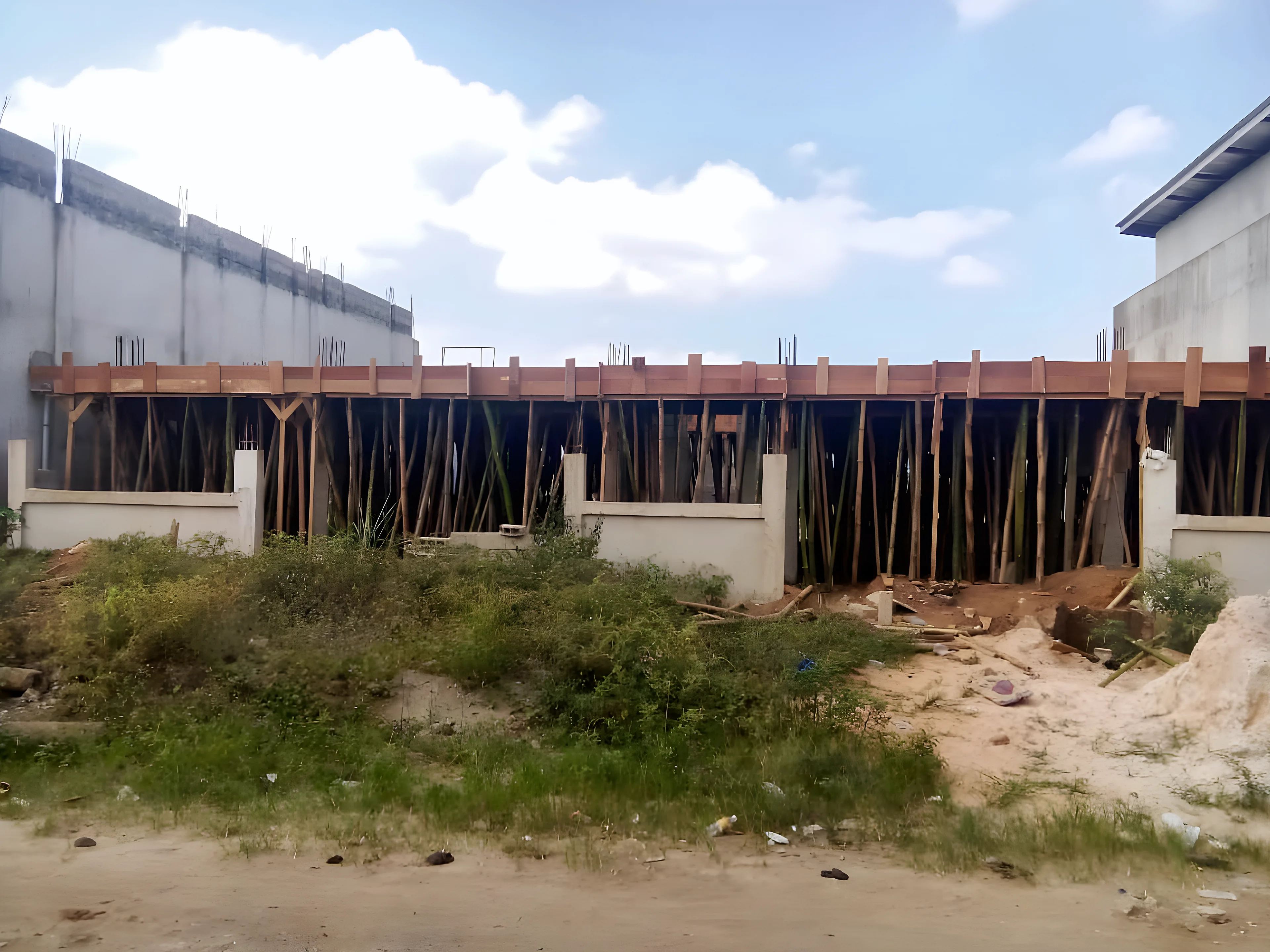 Photo réelle du chantier du Bâtiment A, vue de face : murets du rez-de-chaussée, étais en bambou et coffrage bois de la dalle du 1er étage, attentes d’armatures visibles